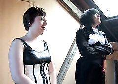 Dominant women in corsets and stockings discipline bare submissives in a rustic wooden dungeon
